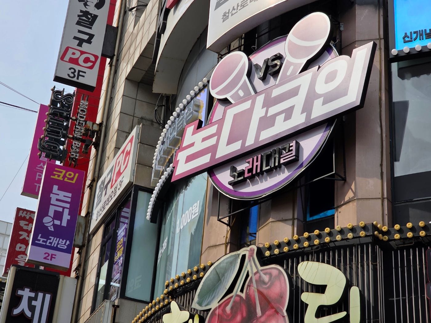 노래방간판, 설치 전에 공장에서 불부터 켜보는 이유가 있을까요? – 비스듬히 올려다본 원형 간판과 건물 주변 거리 모습